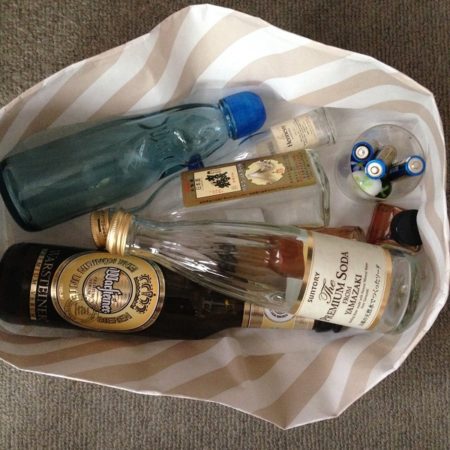 百貨店の紙袋を折り畳んで作った紙箱に、ビールやラムネやリキュールの空瓶が数本保管されている写真。写真は箱の真上から撮影されている。ラムネの水色の空瓶と、SUNTORY PREMIUM SODAの透明な空瓶が写っている。