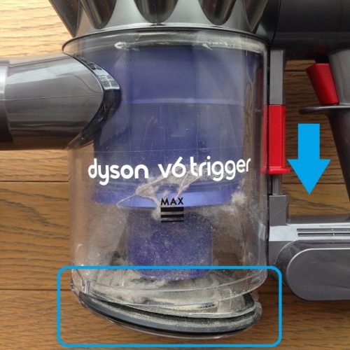 布団掃除機Dyson V6 Triggerのダストトレイを少し開いた状態で撮影した写真。赤いレバーを下向きに1度押すと、透明なダストトレイの底が抜けるようにしてトレイが開く。ダストトレイの中に埃が入っているのが見える