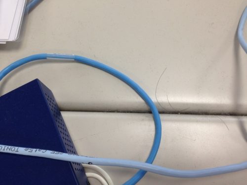 気管支喘息を発症した会社で、当時置かれていた執務机を撮影した写真。薄い灰色の机の上に、はっきり見えるくらい埃が積もっており、髪の毛が2本落ちている。水色と青色の2種類のLANケーブルのコードが、机の上に這っている。