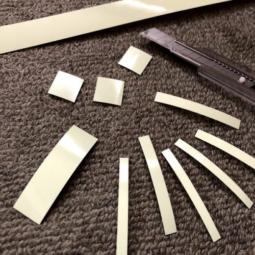 高輝度蓄光テープ「アルファフラッシュ」が、正方形や細長い長方形に切られている写真。写真の右側にはカッターナイフが写っている。