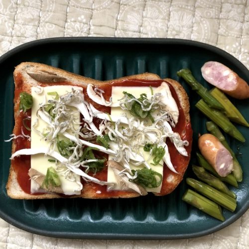 ケチャップを塗りチーズをのせた生のトーストが、不思議なお皿の上に載っている画像。不思議なお皿の上には、短く切ったアスパラガスやソーセージも載せられている。